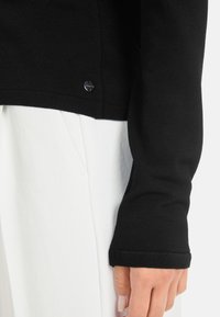 Schwarzer Strickpullover mit langen Ärmeln, mit einem gestickten Saum und einem kleinen metallischen Logo-Detail nahe dem Saum. Kombiniert mit weißen Hosen.