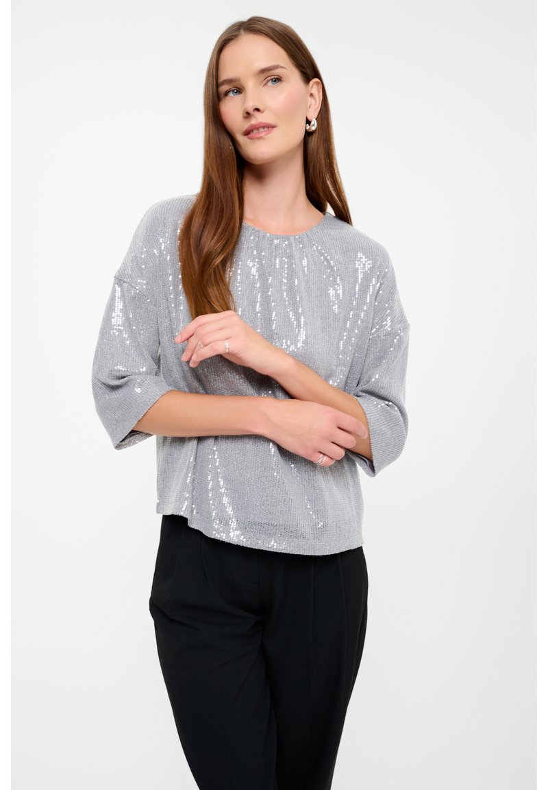 Top grigio con paillettes, scollatura rotonda e vestibilità morbida, con maniche corte e orlo leggermente accorciato. Indossato con pantaloni neri.