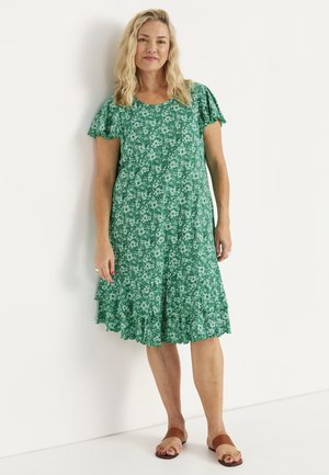 Kvinde med blondt hår iført en kortærmet grøn floral knælængde kjole og brune sandaler, stående op ad en hvid væg.