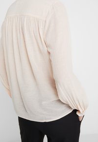 Světle růžová blůza vyrobená z průhledné tkaniny s dlouhými, volnými rukávy a nařasenými detaily na zadní straně, doplněná černými kalhotami.