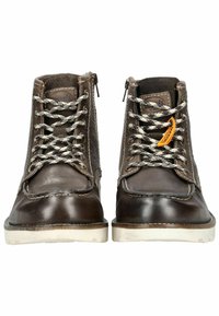 Bottes en cuir marron avec surface texturée, semelles en caoutchouc blanches contrastantes, lacets bicolores et un écusson logo sur le côté.