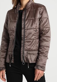 Brązowa puchowa kurtka z zamkiem z przodu, wysokim kołnierzem i elastycznym dołem. Charakteryzuje się teksturowanym wykończeniem i marszczonymi mankietami.