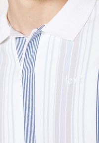 Gros plan d'une personne portant un polo blanc à rayures verticales bleues et grises avec un logo Levi's brodé sur la poitrine.