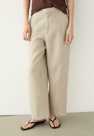 Persona che indossa pantaloni beige a gamba larga e infradito nere, in piedi contro un muro bianco semplice e pavimento grigio chiaro.