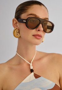 Γυναίκα με μαλλιά χτενισμένα προς τα πίσω, φοράει τεράστια καφέ γυαλιά ηλίου, μεγάλα χρυσά σκουλαρίκια σπειροειδούς σχεδίου και τοπ με λαιμόκοψη halter και διακόσμηση με χάντρες.