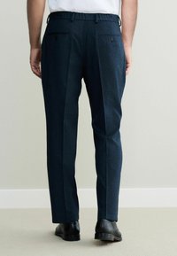 Navy trousers with a smooth texture, featuring two back pockets, a tailored fit, and pleats for added detail, paired with black shoes.