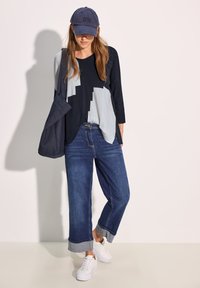 Blaues Top mit drei Viertelärmel und geometrischen grauen Mustern, kombiniert mit dunkelblauen weitgeschnittenen Jeans und weißen Sneakers. Marineblaues Cap getragen.