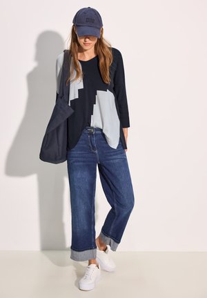 Blaues Top mit drei Viertelärmel und geometrischen grauen Mustern, kombiniert mit dunkelblauen weitgeschnittenen Jeans und weißen Sneakers. Marineblaues Cap getragen.