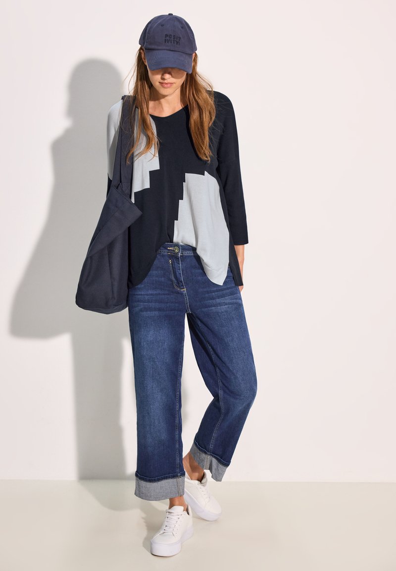 Blaues Top mit drei Viertelärmel und geometrischen grauen Mustern, kombiniert mit dunkelblauen weitgeschnittenen Jeans und weißen Sneakers. Marineblaues Cap getragen.