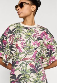 Τοπ με φλοράλ εκτύπωση που απεικονίζει ροζ πουλιά και πράσινα φύλλα σε κρεμ φόντο. Χαλαρή γραμμή με κοντά μανίκια και στρογγυλή λαιμόκοψη.