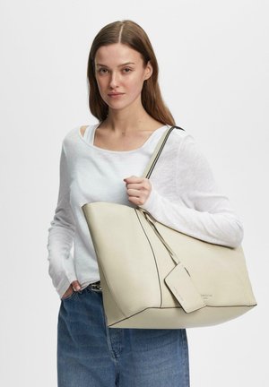 Junge Frau mit langen braunen Haaren trägt ein weißes Langarmshirt und blaue Jeans und hält eine große cremefarbene Leder-Shopper-Tasche auf der Schulter.