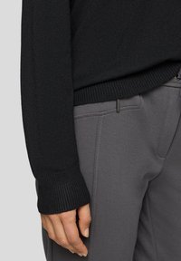 Schwarzer Strickpullover mit gerippten Bündchen, kombiniert mit grauer, strukturierter Hose mit dezentem Taschenakzent.