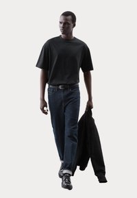 T-shirt nera a maniche corte, jeans in denim blu e scarpe nere. Tiene in mano una giacca nera. Design semplice con vestibilità rilassata e taglio dritto.