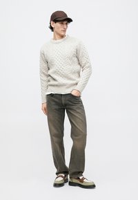 Jeune homme portant un pull en tricot beige, un jean brun délavé, des mocassins bicolores et une casquette de baseball marron, se tenant avec une main dans la poche.