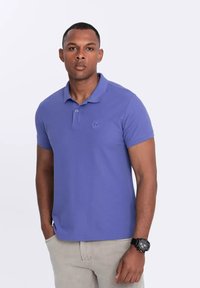 Camisa polo morada con cuello, mangas cortas y un detalle de logo en el pecho. Llevada con pantalones de colores claros y un reloj en la muñeca.