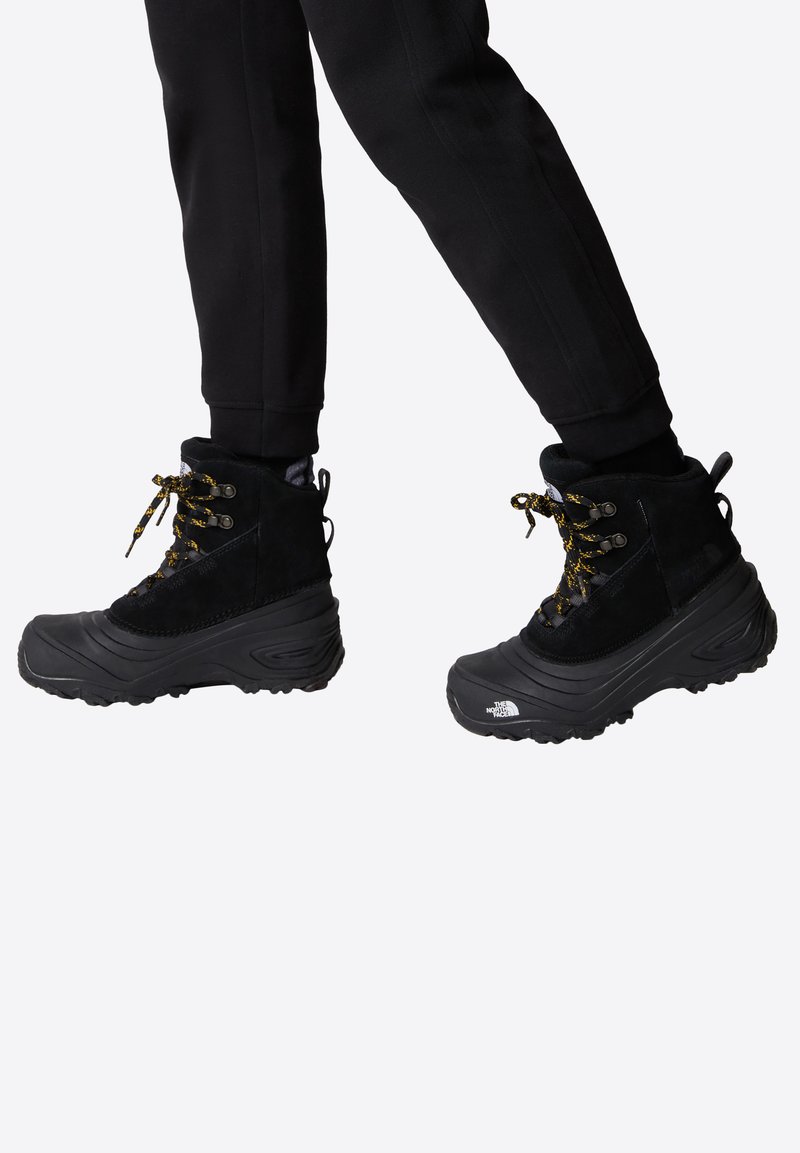 Black waterproof boots with a high ankle, textured suede and rubber materials, featuring yellow laces and a distinct patterned sole.
