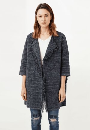 Cárdigan desgastado gris oscuro con mangas anchas y abertura frontal, usado sobre una camiseta blanca y unos jeans azules desgastados con texto en negro.