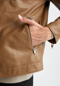 Una mano parzialmente dentro della tasca con zip di una giacca di pelle marrone, indossata sopra un maglione beige e pantaloni neri.