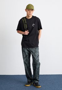Camiseta de algodón negra con logo, pantalones de camuflaje verdes, gorra verde y zapatillas negras con suelas amarillas. Mochila con correas verdes.