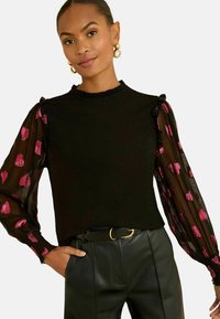 Nő fekete, átlátszó hosszú ujjú, rózsaszín szívmintás felsőben és fekete bőr nadrágban, övvel, az egyik kezét a zsebében tartva pózol.