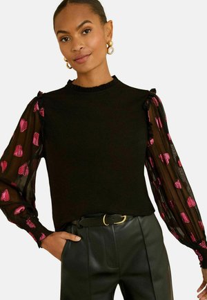 Nő fekete, átlátszó hosszú ujjú, rózsaszín szívmintás felsőben és fekete bőr nadrágban, övvel, az egyik kezét a zsebében tartva pózol.