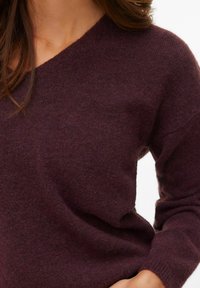 Bordo pleteni džemper s V-izrezom, teksturiranim završetkom i dugim rukavima. Meki materijal s opuštenim krojem i suptilnim detaljima šavova.