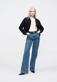 Crna traper jakna preko svijetložute majice, uparena sa širokim plavim trapericama i crnim remenom s velikom kopčom u obliku srca. Glatke teksture.