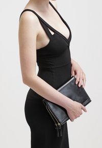 Schwarz geripptes Kleid mit tiefem V-Ausschnitt und breiten Trägern. Das Model hält eine schwarze Ledertasche mit goldfarbenem Reißverschluss und Quastenakzenten.