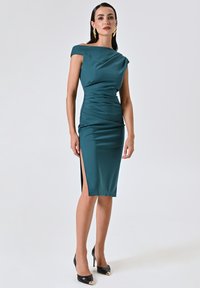 Teal Off-Shoulder-Kleid aus dehnbarem Stoff, mit drapierten Details und einem Oberschenkel-spalt. Kombiniert mit schwarzen Stilettos.