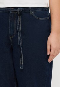 Pantaloni din denim albastru închis cu talie elastică, având cusături galbene, buzunare laterale și o croială lejeră. Textura netedă este evidentă.