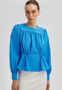 Blusa blu a maniche lunghe con scollatura tonda, corpo arricciato e pieghe sottili. Presenta dettagli con bottoni nella parte anteriore e vita elasticizzata.