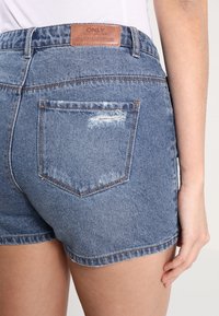 Pantalones cortos de denim en azul medio con dobladillo deshilachado, que cuentan con un parche de cuero de la marca y un bolsillo trasero con detalles desgastados.