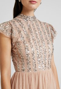 Beige klänning med hög hals och korta, volangärmar. Dekorerad med vertikala rader av silverpaljetter mot ett genomskinligt tyg. Tyllkjol.