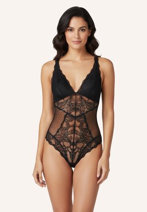 Vrouw staand, voorwaarts gericht, gekleed in een zwarte kanten body met bloemen- en meshpatronen en getande randen aan de bandjes en pijpen.