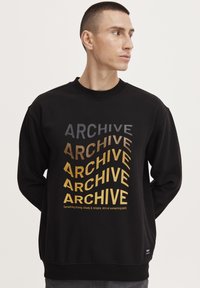 Crna dukserica s velikim, različitim fontovima u sivoj i zlatnoj boji na kojima piše "ARCHIVE." Ribljen ovratnik i manšete, s malim logotipom na rubu.