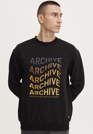 Čierna mikina s veľkými, rôznymi písmami v šedej a zlatej farbe s nápisom "ARCHIVE." Hladký nápletný golier a manžety, s malou logom značky na leme.