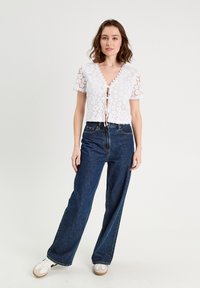 Haut en dentelle blanche avec un col en V et des manches courtes, associé à un jean bleu foncé à taille haute et des baskets blanches.