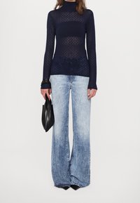 Dolcevita a maglia blu navy con motivo a maglia aperta, abbinato a jeans a zampa di colore azzurro chiaro. Borsa clutch nera tenuta in mano.