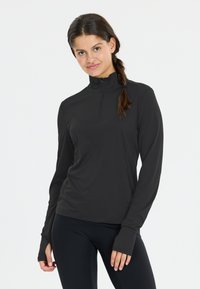 Vrouw met bruin gevlochten haar, gekleed in een zwarte lange mouwen ritssluiting top en zwarte leggings, staande tegen een effen witte achtergrond.
