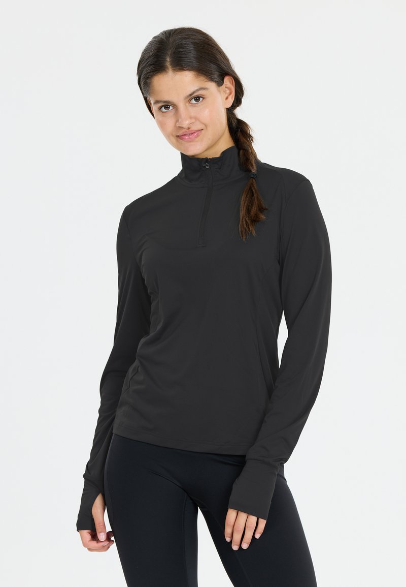 Vrouw met bruin gevlochten haar, gekleed in een zwarte lange mouwen ritssluiting top en zwarte leggings, staande tegen een effen witte achtergrond.
