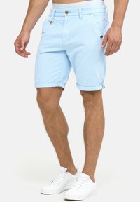 Lyseblå shorts lavet af bomuldsblanding, med optrukne bukseben, knaplukning og sidelommer, båret med hvide sneakers.