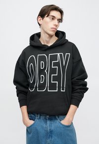 Fekete pamut kapucnis pulóver nagy fehér "OBEY" felirattal. Kenguruzsebbel és húzózsinóros kapucnival rendelkezik. Világoskék farmernadrággal viselve.