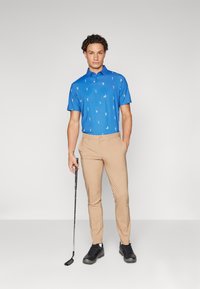 Zils polo krekls ar mazu golfa figūru rakstu, apvienots ar brūnām biksēm. Modelis tur golfa nūju un valkā melnas golfa kurpes.
