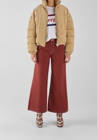 Chaqueta acolchada beige, camiseta gráfica blanca, pantalones anchos color óxido y zapatos de plataforma con estampado a cuadros. Costuras prominentes y textura acolchada.