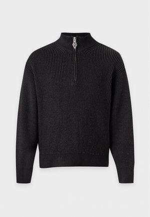 Un mannequin homme se tient devant un fond uni, vêtu d’une casquette noire, d’un pull côtelé à demi-zip, d’un pantalon gris et de baskets noires.