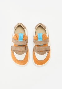 Des chaussures pour tout-petits avec une combinaison de daim orange, crème et marron, dotées de deux bandes Velcro réglables et d'une étiquette de marque bleue.
