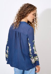 Marineblaue Bluse mit langen Ärmeln, die mit floralem Stickerei in Pink und Grün an den Ärmeln verziert ist. Leichter Stoff und lässige Passform.