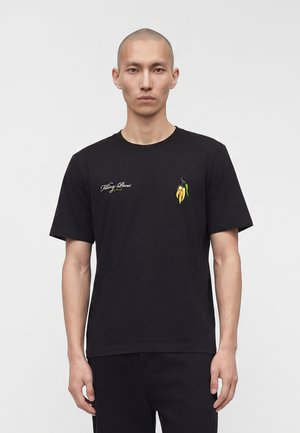 Borotvált fejű férfi, fekete pólót visel, melyen sárga chili paprika grafika és a „Filling Pieces” felirat látható a mellkasán, egyszínű fehér háttér előtt áll.