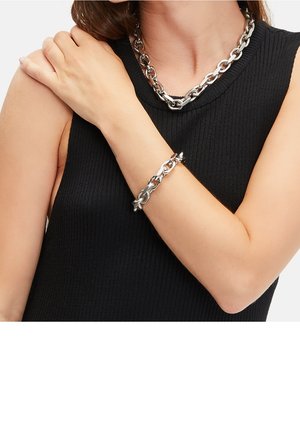 Femme portant un haut noir sans manches avec un collier chaîne argenté épais et un bracelet assorti, la main reposant sur l'épaule opposée.