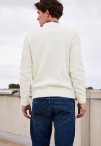 Cremfarbener Strickpullover mit einem strukturierten Muster, kombiniert mit dunkelblauen Jeans. Der Pullover hat gerippte Bündchen und einen gerippten Saum.